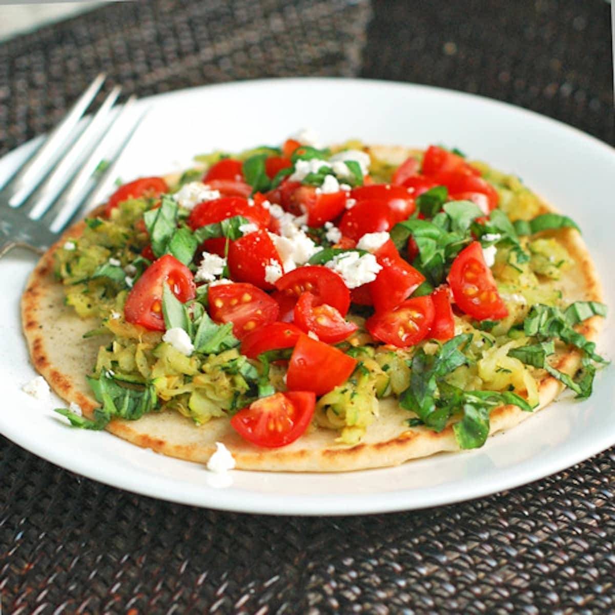 Flatbread pizza with zucchini, cherry tomatoes, and basil on a plate with a fork.